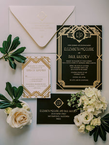 Photo of deco invitation suite from above. Black invitation with gold foil, black reply envelope with gold foil, white reply card with deco design in gold foil. White flowers and green leaves laying next to the invitation.